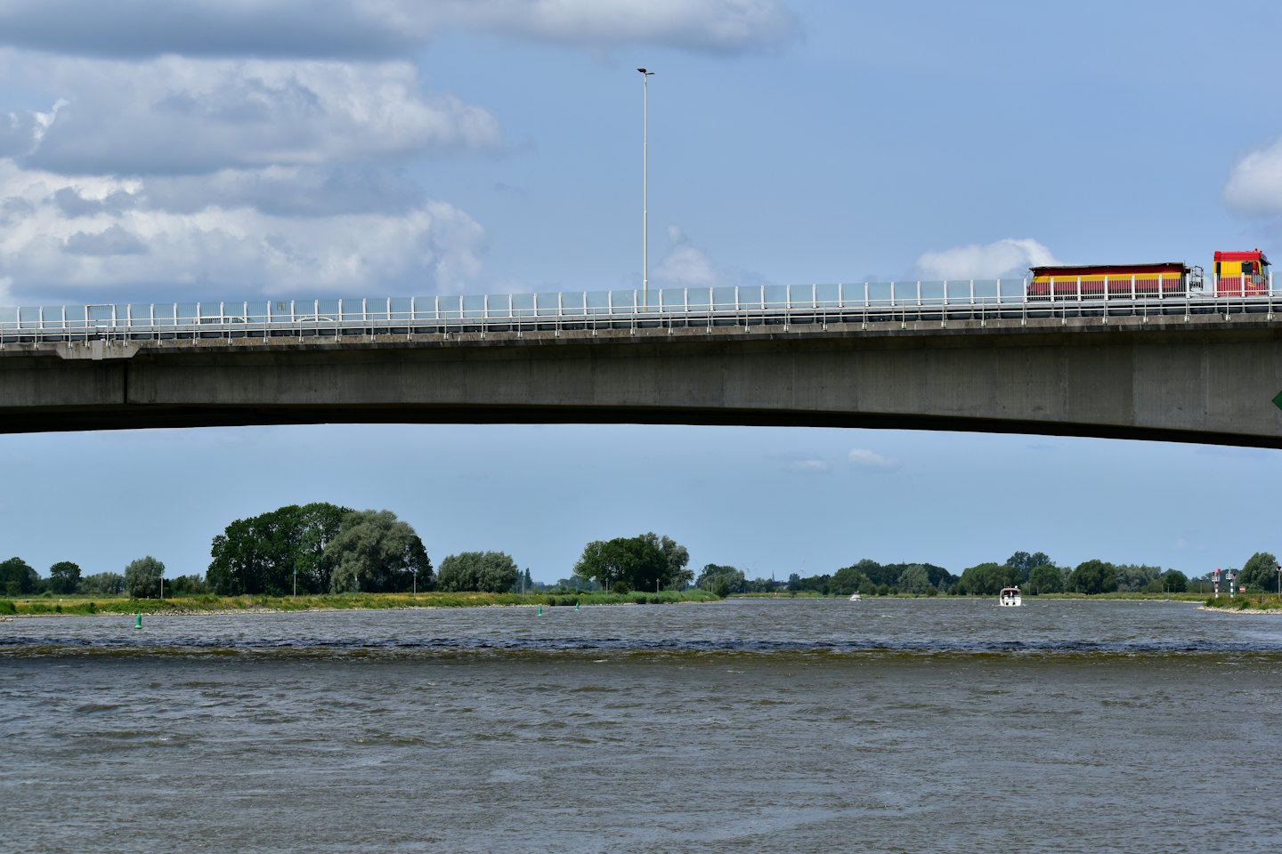 A red and yellow semi-truck drives over a concrete bridge crossing a wide river.